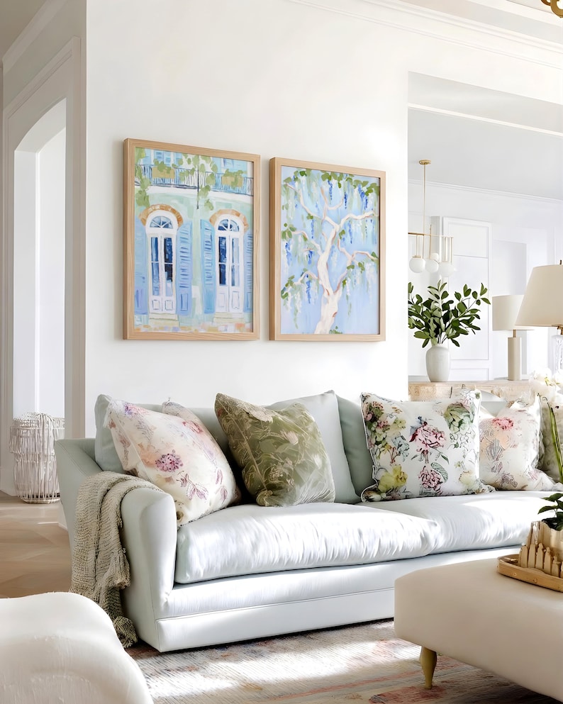 Puede incluir: Un sof&aacute; azul claro con cojines florales y estampados y una manta beige. Dos pinturas enmarcadas cuelgan sobre el sof&aacute;, una que representa un edificio con contraventanas azules y la otra un &aacute;rbol con flores colgantes. La habitaci&oacute;n tiene un ambiente luminoso y aireado.