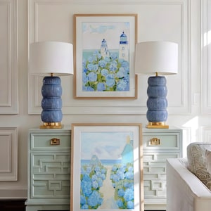 May include: Two blue ceramic lamps with white shades sit atop matching light blue dressers. Artwork featuring lighthouses and a coastal path with blue flowers hangs above the dressers. The room has white paneling.