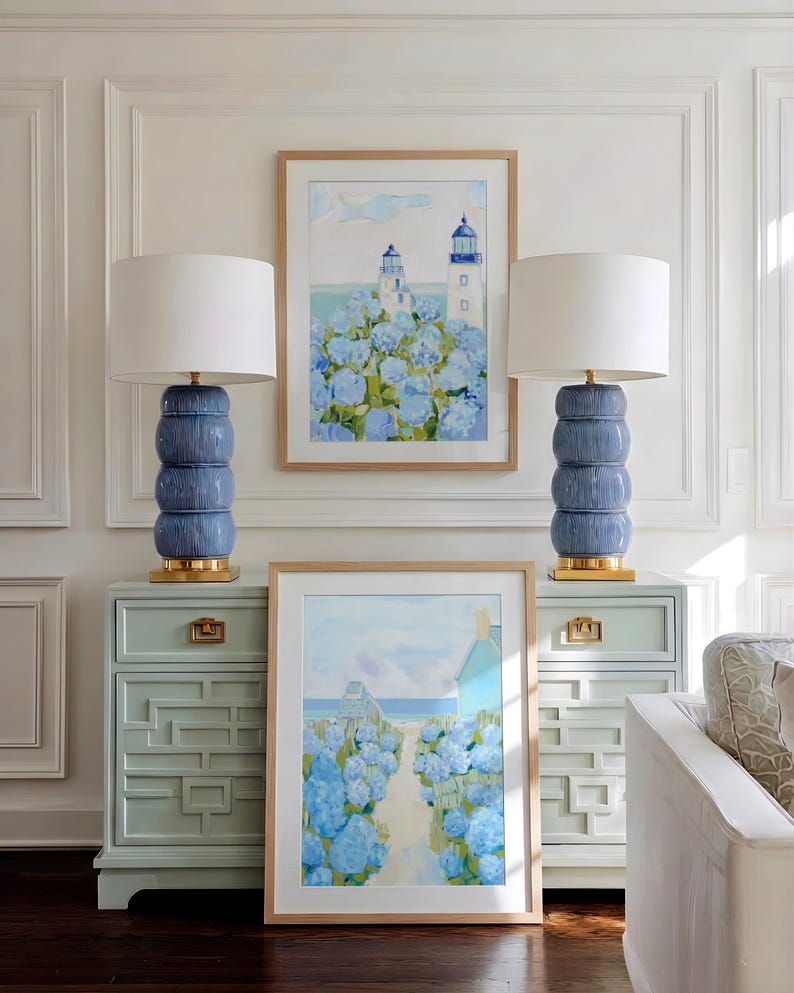 May include: Two blue ceramic lamps with white shades sit atop matching light blue dressers. Artwork featuring lighthouses and a coastal path with blue flowers hangs above the dressers. The room has white paneling.