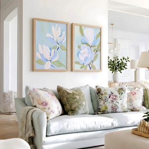 May include: A light blue sofa with several decorative pillows in various floral patterns. Two framed paintings of white and blue flowers hang above the sofa. A neutral-toned throw blanket is draped over the arm of the sofa.
