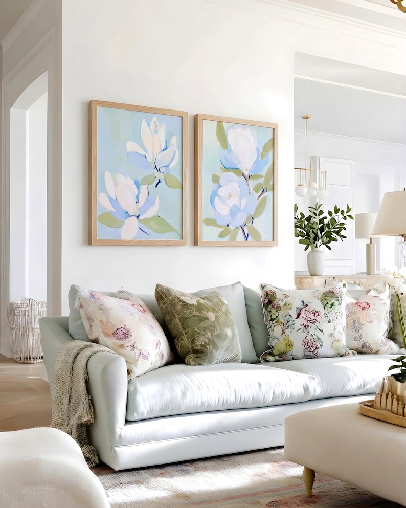 May include: A light blue sofa with several decorative pillows in various floral patterns. Two framed paintings of white and blue flowers hang above the sofa. A neutral-toned throw blanket is draped over the arm of the sofa.