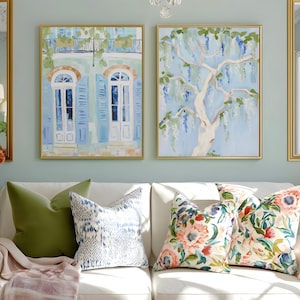 Puede incluir: Dos pinturas enmarcadas con marcos dorados cuelgan sobre un sof&aacute; blanco con cojines decorativos. Una pintura representa un edificio con contraventanas azules y puertas blancas. La otra muestra un &aacute;rbol con flores moradas colgantes.