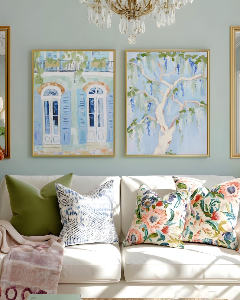 Puede incluir: Dos pinturas enmarcadas con marcos dorados cuelgan sobre un sof&aacute; blanco con cojines decorativos. Una pintura representa un edificio con contraventanas azules y puertas blancas. La otra muestra un &aacute;rbol con flores moradas colgantes.