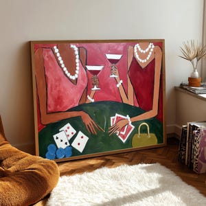 May include: An abstract painting depicts two figures in red dresses, holding martini glasses. They are seated at a green table with playing cards and a small yellow purse. The artwork is framed in a wooden frame and leans against a wall.