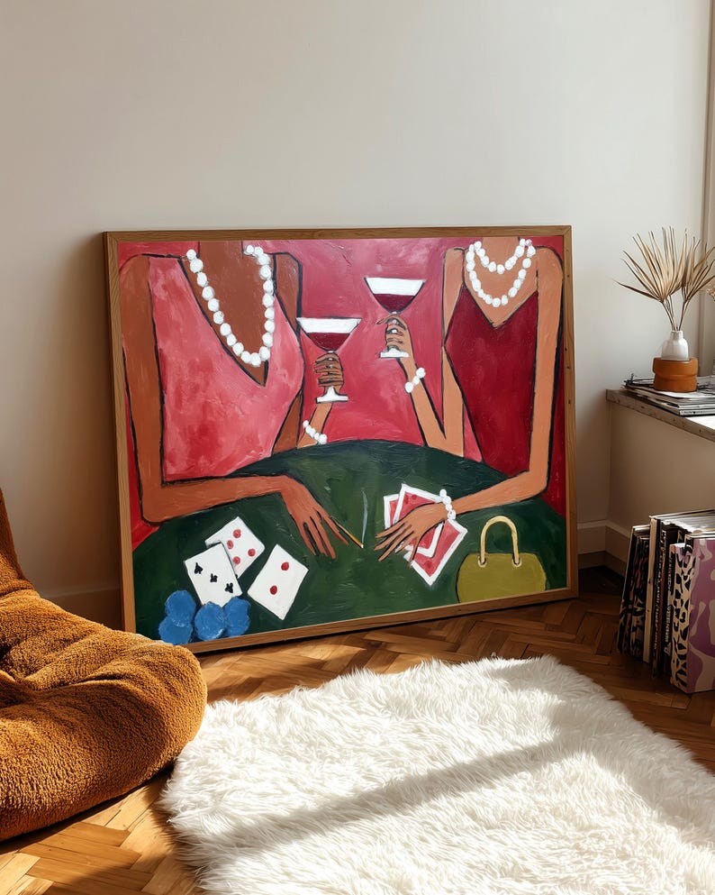 May include: An abstract painting depicts two figures in red dresses, holding martini glasses. They are seated at a green table with playing cards and a small yellow purse. The artwork is framed in a wooden frame and leans against a wall.