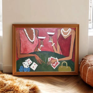 May include: A framed painting depicts two figures at a table playing cards. The figures wear pink and red dresses and pearl necklaces. A green table holds cards, blue chips, and drinks. The artwork is in a wooden frame.
