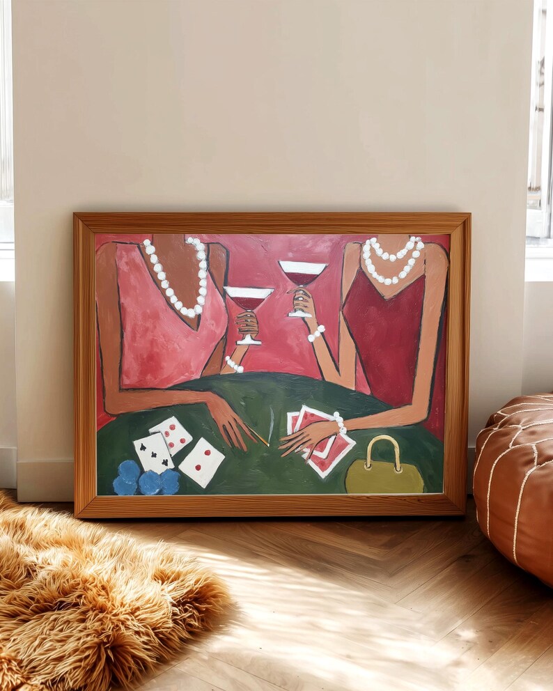 May include: A framed painting depicts two figures at a table playing cards. The figures wear pink and red dresses and pearl necklaces. A green table holds cards, blue chips, and drinks. The artwork is in a wooden frame.