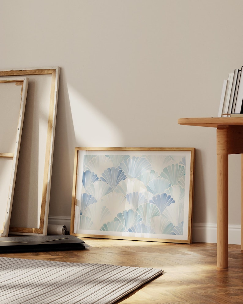 May include: A framed print with a blue and white watercolor pattern of overlapping fan shapes. The print is hanging on a white wall in a room with wooden floors and a wooden table.