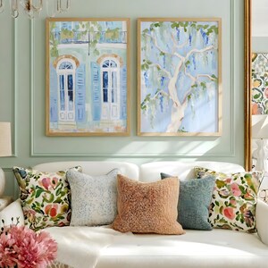 Puede incluir: Dos pinturas en acuarela enmarcadas cuelgan sobre un sof&aacute; blanco con cojines decorativos. Una pintura representa un balc&oacute;n con contraventanas y puertas azules. La otra muestra un &aacute;rbol con flores moradas en cascada. La habitaci&oacute;n tiene una pared verde claro.