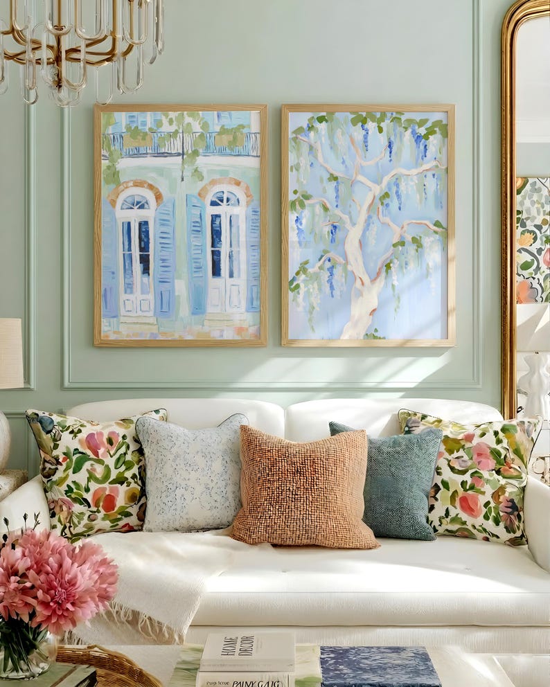 Puede incluir: Dos pinturas en acuarela enmarcadas cuelgan sobre un sof&aacute; blanco con cojines decorativos. Una pintura representa un balc&oacute;n con contraventanas y puertas azules. La otra muestra un &aacute;rbol con flores moradas en cascada. La habitaci&oacute;n tiene una pared verde claro.