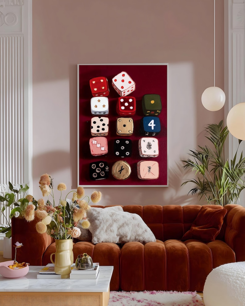 Op de afbeelding: Een ingelijst kunstwerk met twaalf decoratieve dobbelstenen in verschillende kleuren, waaronder rood, wit, zwart en roze, tegen een dieprode achtergrond. De dobbelstenen hebben verschillende patronen en cijfers. Het kunstwerk is boven een bruine bank geplaatst.