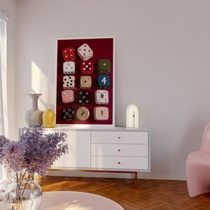 Op de afbeelding: Een ingelijste kunstdruk met twaalf dobbelstenen in verschillende kleuren, waaronder rood, wit, groen en roze, tegen een dieprode achtergrond. De dobbelstenen tonen verschillende punt- en cijfercombinaties. Het kunstwerk wordt in een moderne woonkamer gepresenteerd.