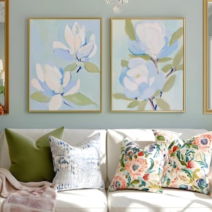 May include: Two framed floral paintings with gold frames hang above a white sofa with decorative pillows. The paintings feature large, abstract white and blue flowers on a light blue background. The sofa has green, patterned, and floral pillows.