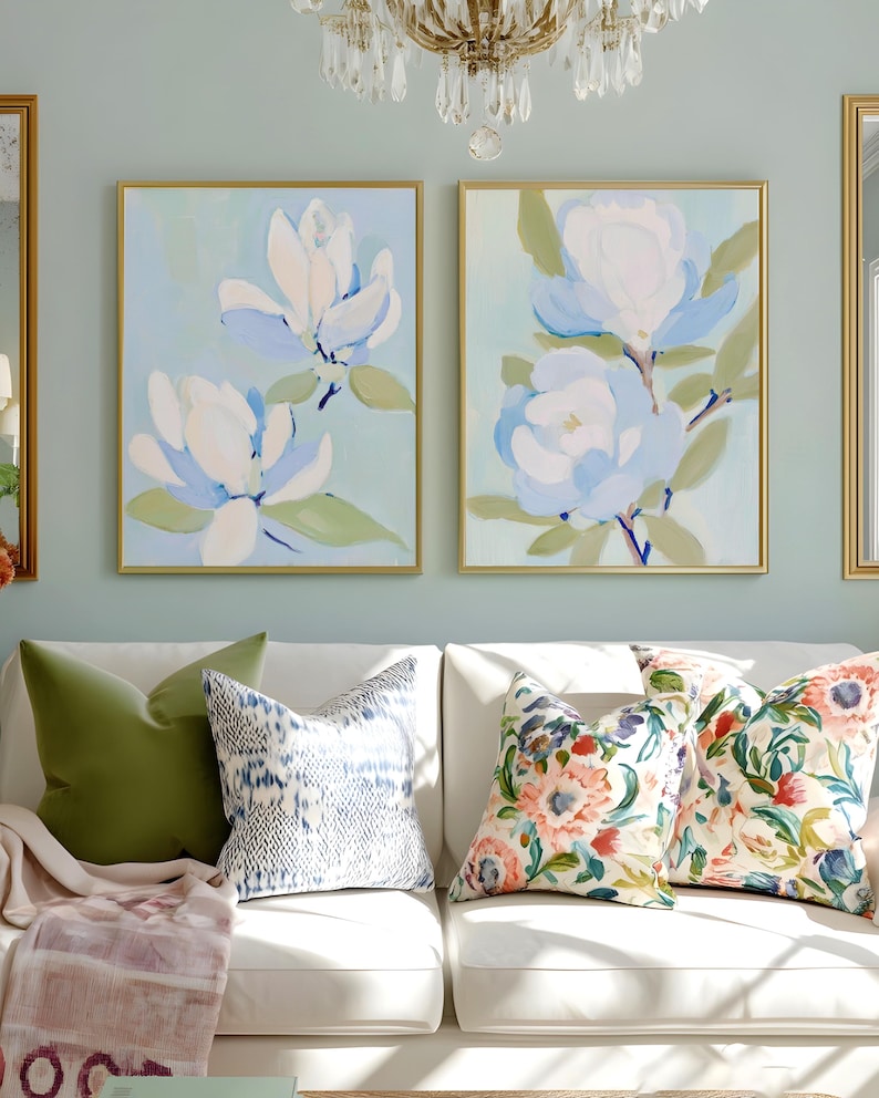 May include: Two framed floral paintings with gold frames hang above a white sofa with decorative pillows. The paintings feature large, abstract white and blue flowers on a light blue background. The sofa has green, patterned, and floral pillows.