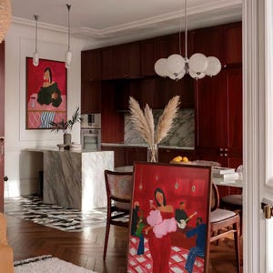 May include: An interior shot of a room with a framed painting leaning against a wall. The painting depicts a woman in a red dress with a pink boa, holding a glass of wine. The room has a marble island, dark wood cabinets, and a patterned rug.