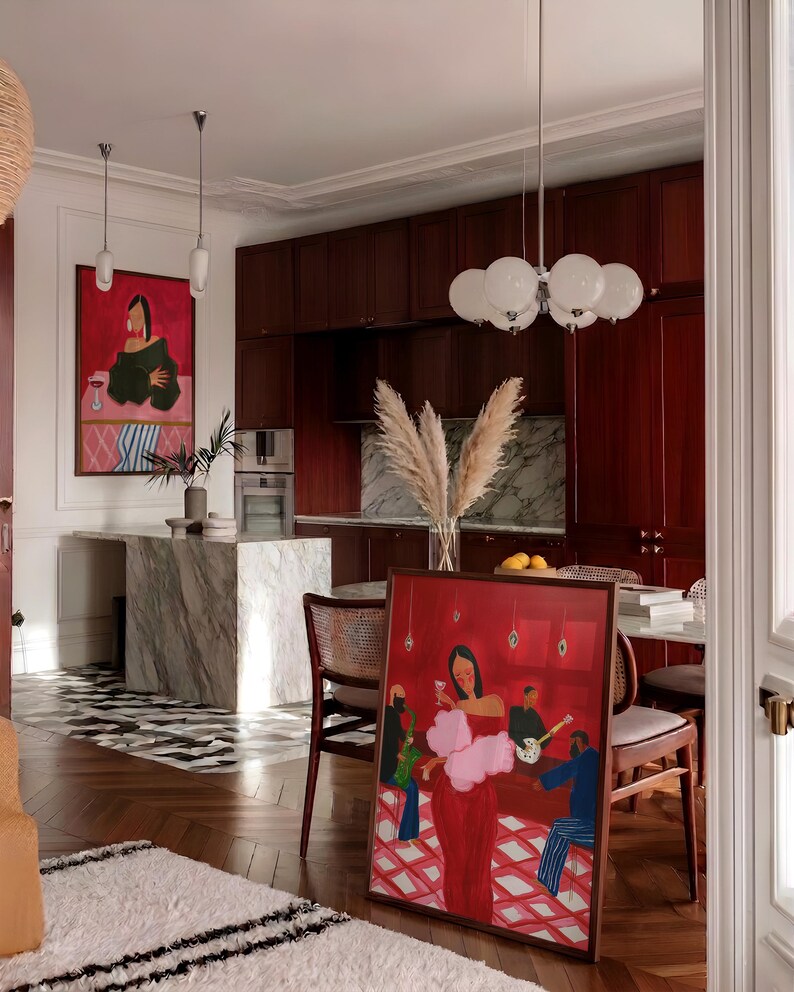 May include: An interior shot of a room with a framed painting leaning against a wall. The painting depicts a woman in a red dress with a pink boa, holding a glass of wine. The room has a marble island, dark wood cabinets, and a patterned rug.