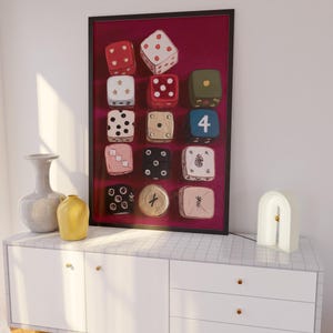 Op de afbeelding: Een ingelijste print met twaalf kleurrijke dobbelstenen tegen een bordeauxrode achtergrond. De dobbelstenen hebben verschillende kleuren en patronen, waaronder rood met witte stippen en een blauwe dobbelsteen met het nummer 4. Het kunstwerk staat boven een witte kast.