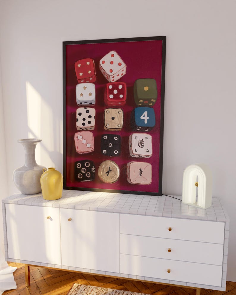 Op de afbeelding: Een ingelijste print met twaalf kleurrijke dobbelstenen tegen een bordeauxrode achtergrond. De dobbelstenen hebben verschillende kleuren en patronen, waaronder rood met witte stippen en een blauwe dobbelsteen met het nummer 4. Het kunstwerk staat boven een witte kast.