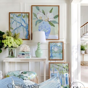 May include: A gallery wall featuring framed art prints with botanical and landscape themes. The artwork includes a large floral painting, a tree scene, and a house with a blue and white color scheme. A white console table holds a lamp and decorative items.
