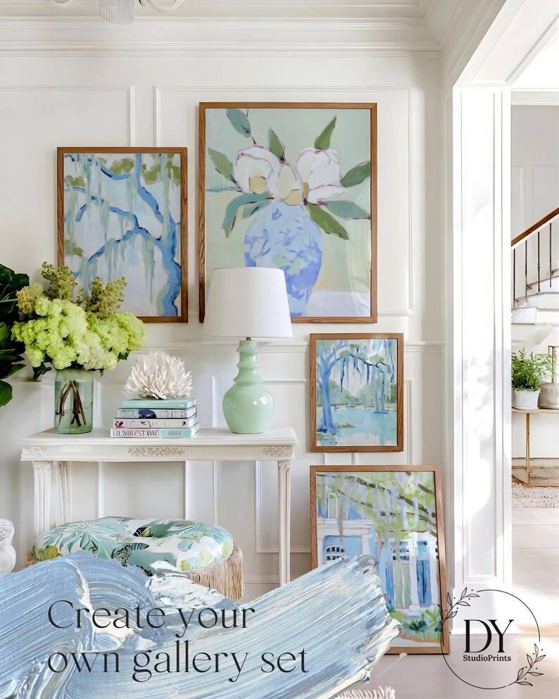 May include: A gallery wall featuring framed art prints with botanical and landscape themes. The artwork includes a large floral painting, a tree scene, and a house with a blue and white color scheme. A white console table holds a lamp and decorative items.