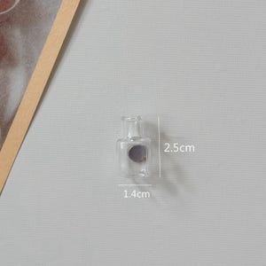 K&ouml;nnte beinhalten: Kleine, transparente Glasflasche mit rundem K&ouml;rper und schmalem Hals. Die Flasche ist 1,4 cm breit und 2,5 cm hoch. Auf der Vorderseite befindet sich ein graues Etikett. Der Hintergrund ist hellgrau.