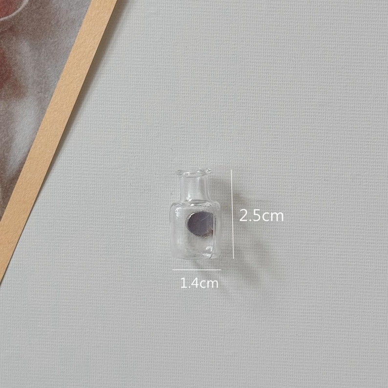 K&ouml;nnte beinhalten: Kleine, transparente Glasflasche mit rundem K&ouml;rper und schmalem Hals. Die Flasche ist 1,4 cm breit und 2,5 cm hoch. Auf der Vorderseite befindet sich ein graues Etikett. Der Hintergrund ist hellgrau.