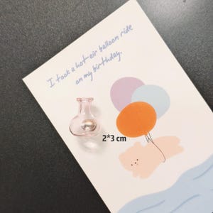 K&ouml;nnte beinhalten: Ein kleines, rosa Glasfl&auml;schchen mit einer silbernen Kugel im Inneren, neben einer Karte mit einer Hei&szlig;luftballon-Illustration. Auf der Karte steht "I took a hot-air balloon ride on my birthday." Das Fl&auml;schchen misst ca. 2 x 3 cm.