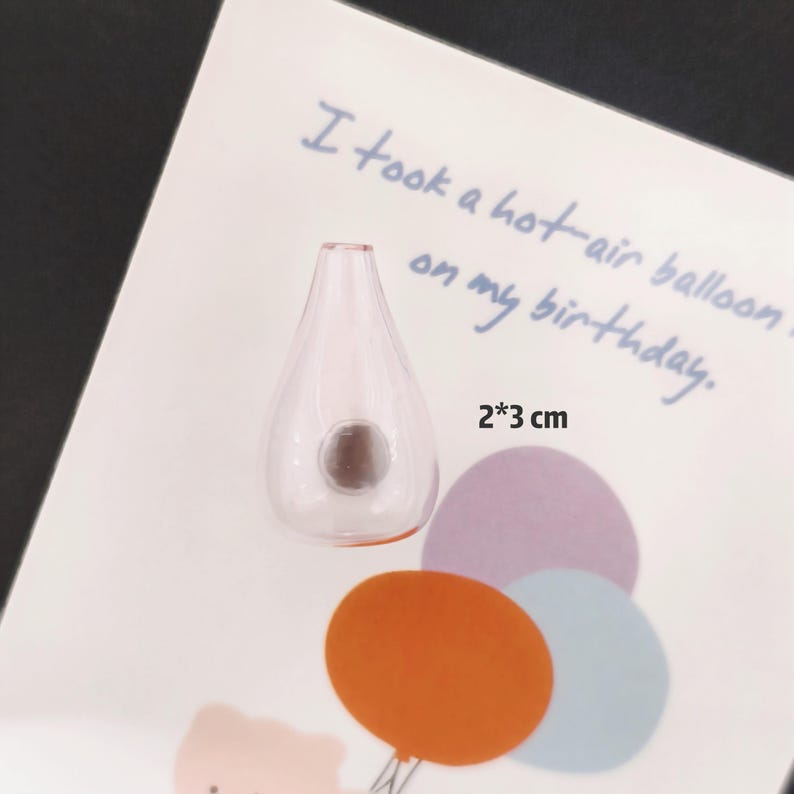 K&ouml;nnte beinhalten: Eine kleine, tropfenf&ouml;rmige Glasflasche mit einem dunklen, braunen Verschluss. Die Flasche misst 2 cm x 3 cm. Das Bild enth&auml;lt den Text "I took a hot-air balloon on my birthday."