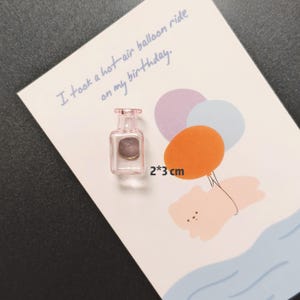 K&ouml;nnte beinhalten: Ein kleiner, rosa Glasmagnet in Flaschenform, 2 x 3 cm, liegt auf einer Karte mit einer Hei&szlig;luftballon-Illustration. Auf der Karte steht handschriftlich: "I took a hot-air balloon ride on my birthday."