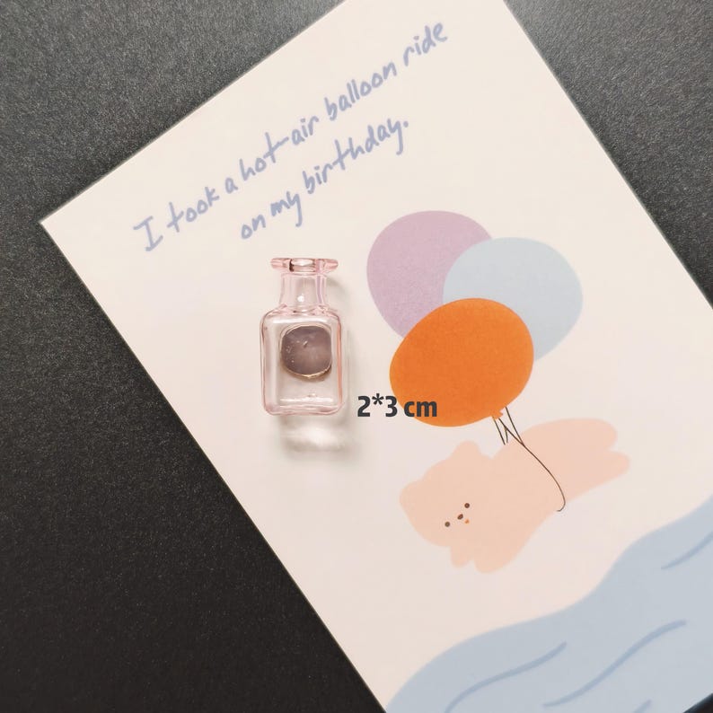 K&ouml;nnte beinhalten: Ein kleiner, rosa Glasmagnet in Flaschenform, 2 x 3 cm, liegt auf einer Karte mit einer Hei&szlig;luftballon-Illustration. Auf der Karte steht handschriftlich: "I took a hot-air balloon ride on my birthday."