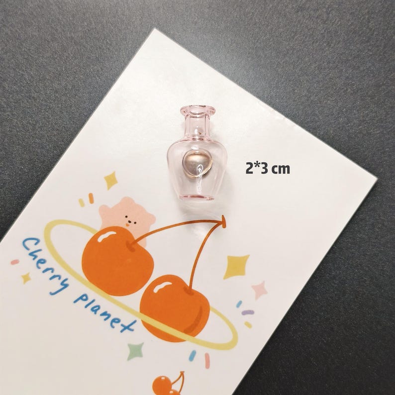 K&ouml;nnte beinhalten: Eine kleine, rosa Glasparf&uuml;mflasche mit silberfarbenem Verschluss, misst 2 x 3 cm. Die Flasche befindet sich auf einer wei&szlig;en Karte mit einem Cartoon-Kirschdesign und dem Schriftzug "Cherry planet".