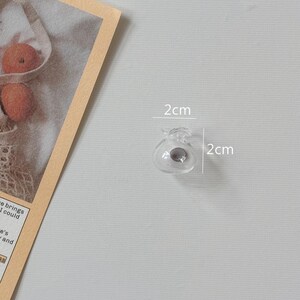 K&ouml;nnte beinhalten: Eine kleine, klare Glasperle, etwa 2 cm im Durchmesser, mit einem dunkelgrauen Zentrum. Die Perle hat eine einzigartige, dekorative Form. Das Bild ist vor einem hellgrauen Hintergrund aufgenommen.