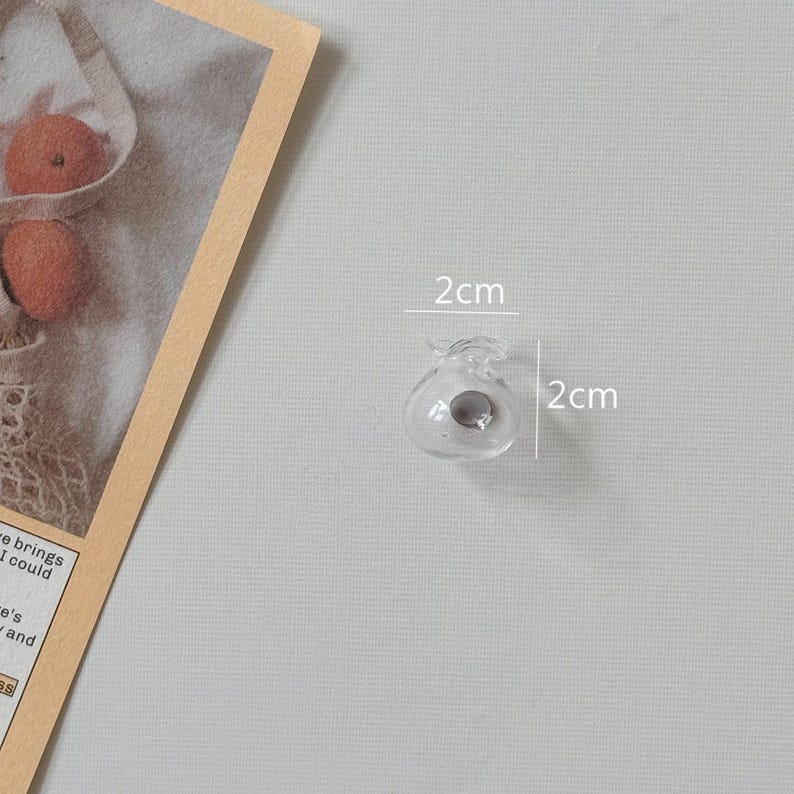 K&ouml;nnte beinhalten: Eine kleine, klare Glasperle, etwa 2 cm im Durchmesser, mit einem dunkelgrauen Zentrum. Die Perle hat eine einzigartige, dekorative Form. Das Bild ist vor einem hellgrauen Hintergrund aufgenommen.