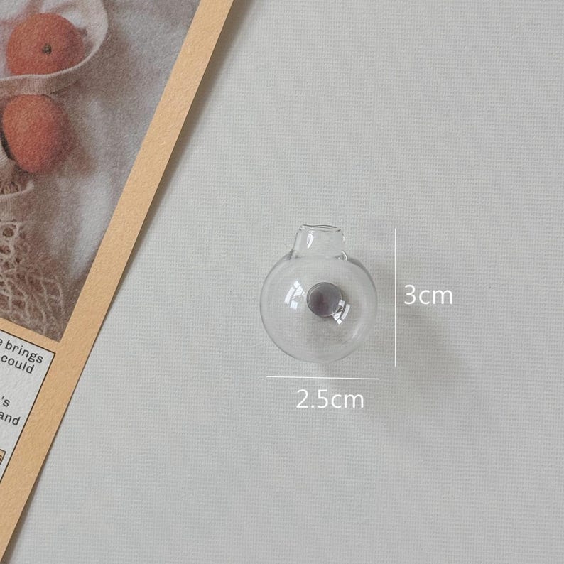 K&ouml;nnte beinhalten: Eine klare, kugelf&ouml;rmige Vase aus Glas mit einer kleinen &Ouml;ffnung oben. Die Vase ist 2,5 cm breit und 3 cm hoch. Im Inneren der Vase ist ein kleines, dunkelgraues Objekt zu sehen. Die Vase steht auf einer hellgrauen Oberfl&auml;che.