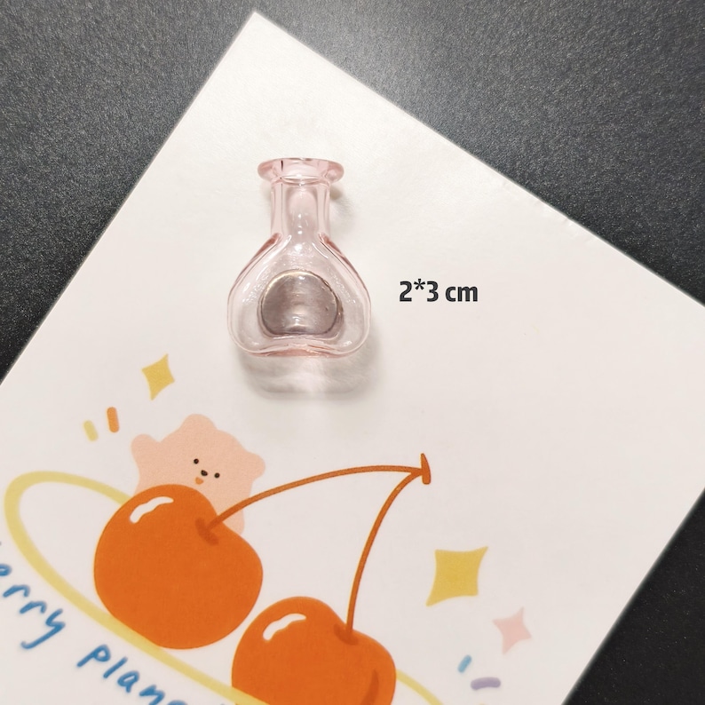 K&ouml;nnte beinhalten: Eine kleine, rosa Glasflasche, ca. 2 cm x 3 cm, liegt auf einer wei&szlig;en Karte mit einer Kirschillustration. Die Flasche hat eine einzigartige Form mit rundem Boden und schmalem Hals. Auf der Karte steht auch der Text "cherry plan".