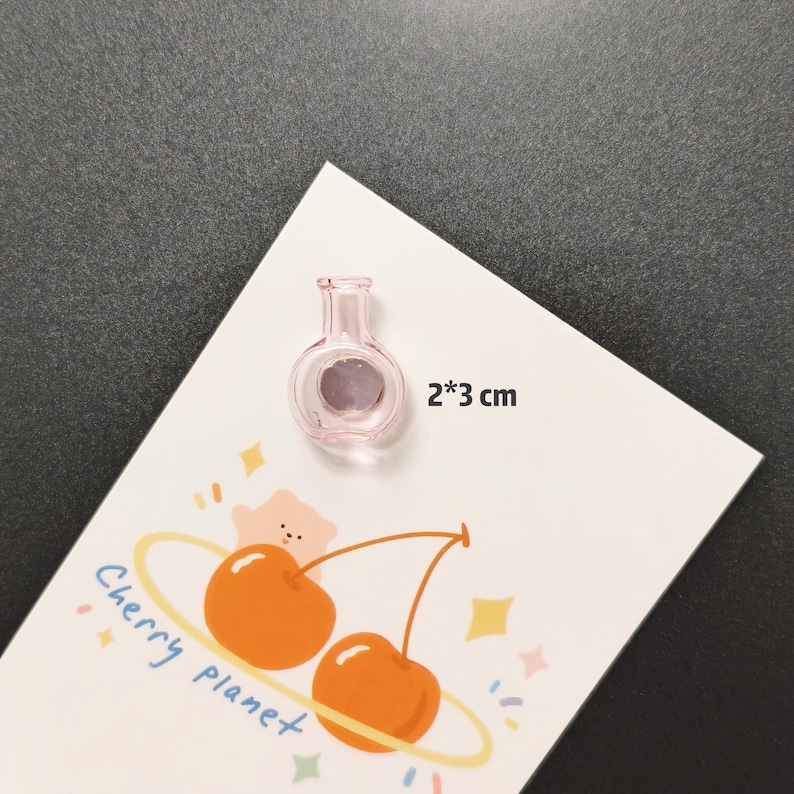 K&ouml;nnte beinhalten: Ein kleiner, rosa Magnet in Flaschenform aus Glas, 2 x 3 cm gro&szlig;, liegt auf einer wei&szlig;en Karte mit einem Kirschdesign. Die Karte zeigt die Worte "Cherry planet" und Illustrationen von Kirschen und Sternen.
