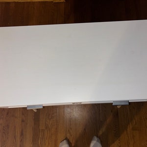 May include: A white rectangular table with a smooth surface. The table has a simple design and is supported by small legs with metal brackets. The table is set on a wooden floor.