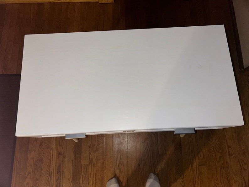 May include: A white rectangular table with a smooth surface. The table has a simple design and is supported by small legs with metal brackets. The table is set on a wooden floor.