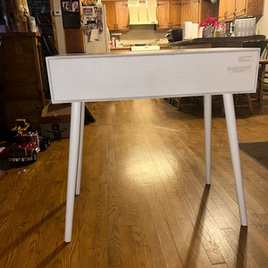 May include: A white rectangular table with four white, angled legs. The table is made of wood and has a simple, modern design. The legs are evenly spaced and provide a sturdy base. The table is suitable for various uses.