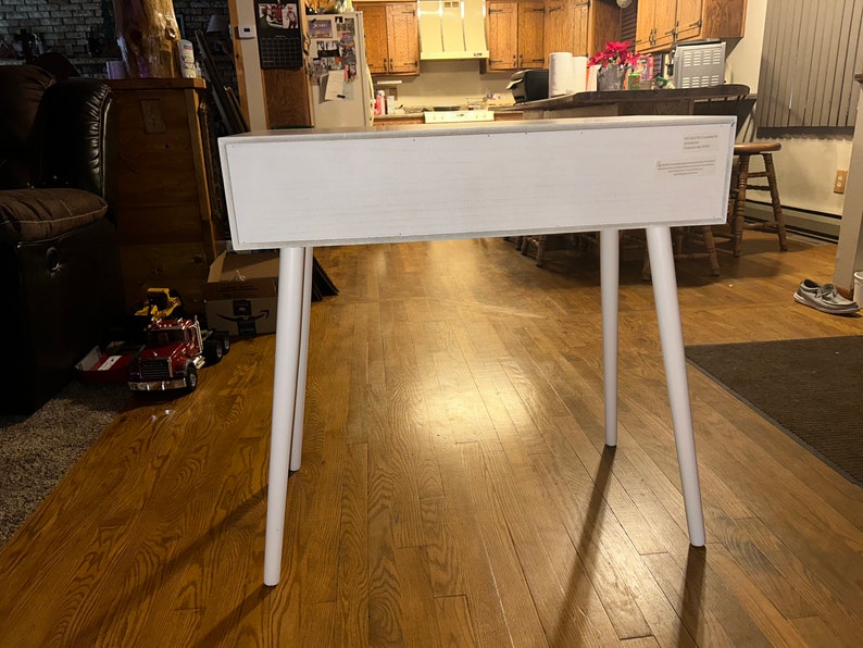 May include: A white rectangular table with four white, angled legs. The table is made of wood and has a simple, modern design. The legs are evenly spaced and provide a sturdy base. The table is suitable for various uses.