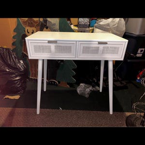May include: White desk with two drawers and a woven cane design on the drawer fronts. The desk has four white, tapered legs. The desk is a light colour and appears to be made of wood. The desk is a modern style.