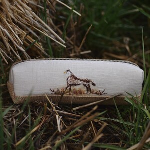 May include: A rectangular pencil case with a beige linen exterior and a burlap base. The case features an embroidered image of a brown and white bird. The pencil case is surrounded by dry grass and green grass.