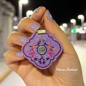 以下が含まれることがあります： ピンクとオレンジの花柄刺繍が施された紫色のフェルトイヤリング。 イヤリングの中央には小さな丸い鏡があり、金色の金属製のフックが付いています。 「Venus Shabani」の文字が見えます。