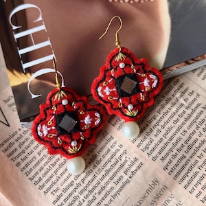 May include: Red and black embroidered earrings with a geometric design and a pearl bead. The earrings have gold-colored hooks and a central square mirror. These unique earrings are a statement piece.