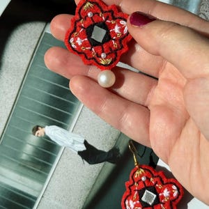 May include: Red and black embroidered earrings with a geometric design, featuring white and gold accents, a small mirror, and a dangling pearl. The earrings have gold-colored hooks.