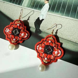 May include: A pair of red felt earrings with a black center and white and gold embroidery. Each earring has a large white pearl at the bottom and gold-colored ear hooks. The earrings are in a decorative, ornate shape.