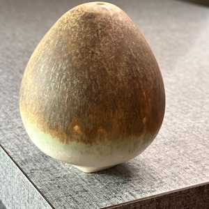 Puede incluir: Un pequeño objeto de cerámica con forma de huevo, parte superior redondeada y base estrecha. La superficie presenta un esmalte moteado en tonos marrón y bronce en la parte superior, que se degrada hacia un color blanco crema en la base.