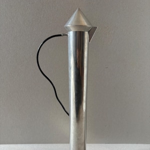 Peut inclure: Une verseuse en métal argenté, de forme élancée, avec un sommet conique et une poignée noire. Le corps est cylindrique et la base évasée. Le fond est gris neutre.