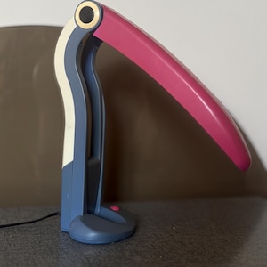 Peut inclure: Lampe de bureau moderne au design inspiré du toucan. La lampe présente un abat-jour incurvé de couleur magenta et un corps bleu, blanc et gris. Un petit bouton rond rose est sur la base. La lampe est branchée sur une prise électrique.