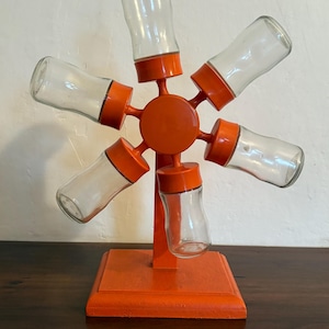 Puede incluir: Estante de especias naranja con seis tarros de vidrio. El estante tiene forma de estrella y tiene una base.
